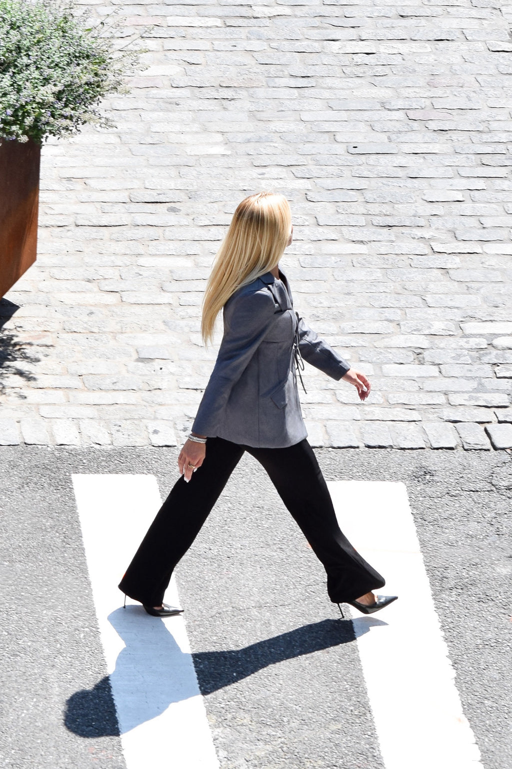 A woman with long blonde hair walks across a white zebra crossing on a paved street, wearing a gray blazer, black wide-leg trousers, and black pointed-toe heels.