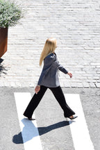 A woman with long blonde hair walks across a white zebra crossing on a paved street, wearing a gray blazer, black wide-leg trousers, and black pointed-toe heels.