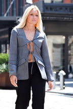 A blonde woman wearing a gray wrap-style blazer with a front tie detail and black pants, standing outdoors on a city street.