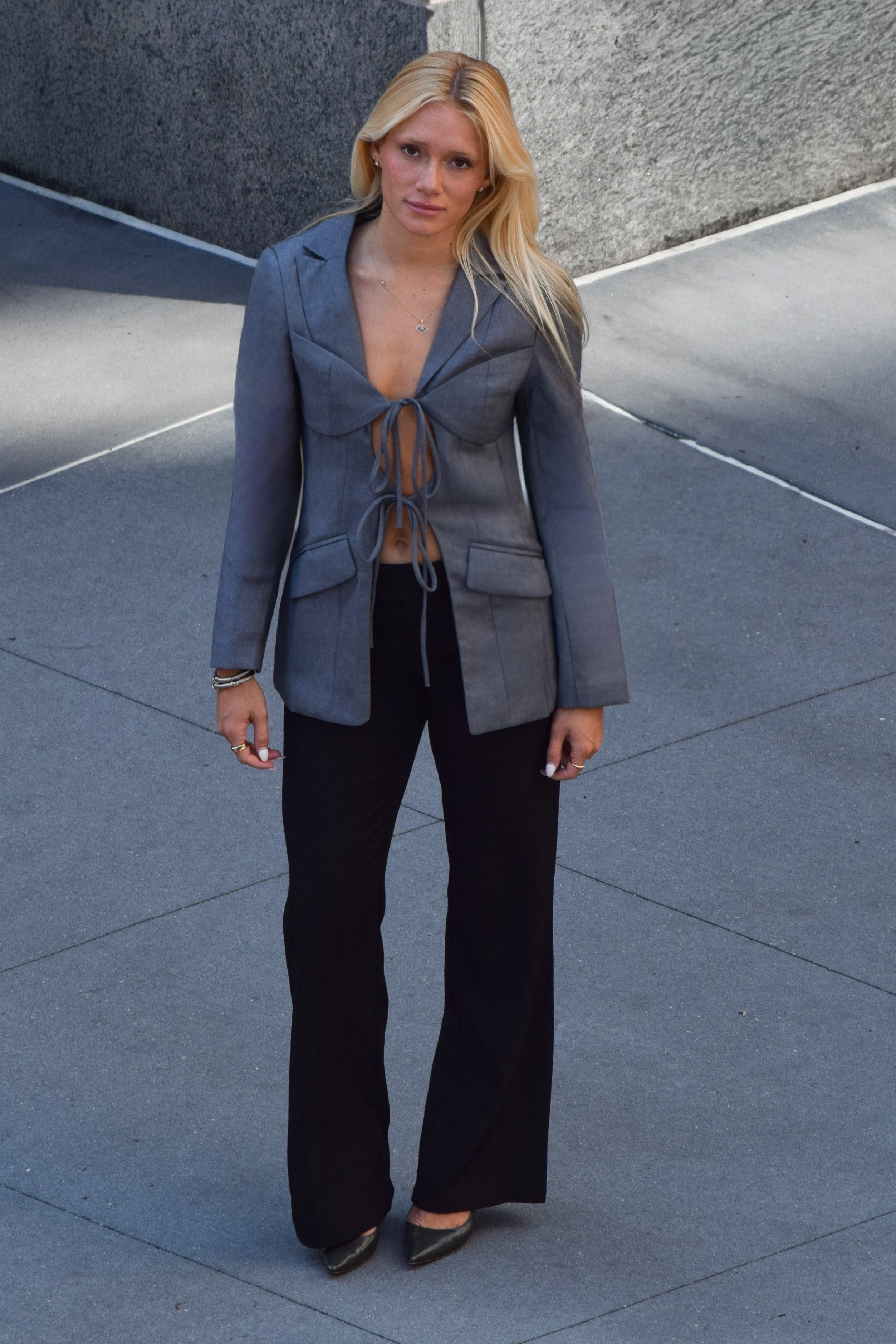 A woman stands outdoors on a paved surface wearing a gray wrap-style blazer with front tie details, black wide-leg pants, and dark pointed flats.