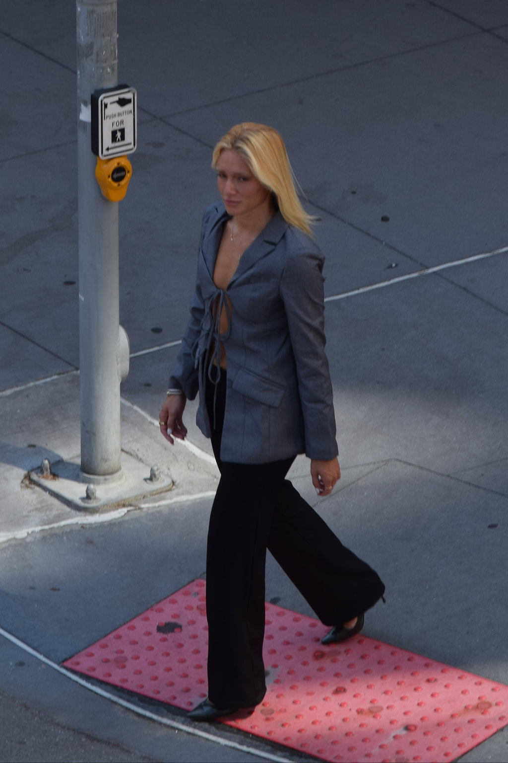 A woman with blonde hair walks across a pink patterned mat on a city sidewalk, wearing a gray blazer and black pants.