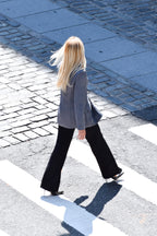A woman with long blonde hair walks across a zebra crossing from behind, wearing a gray blazer, black flared pants, and black heels on a cobblestone street.