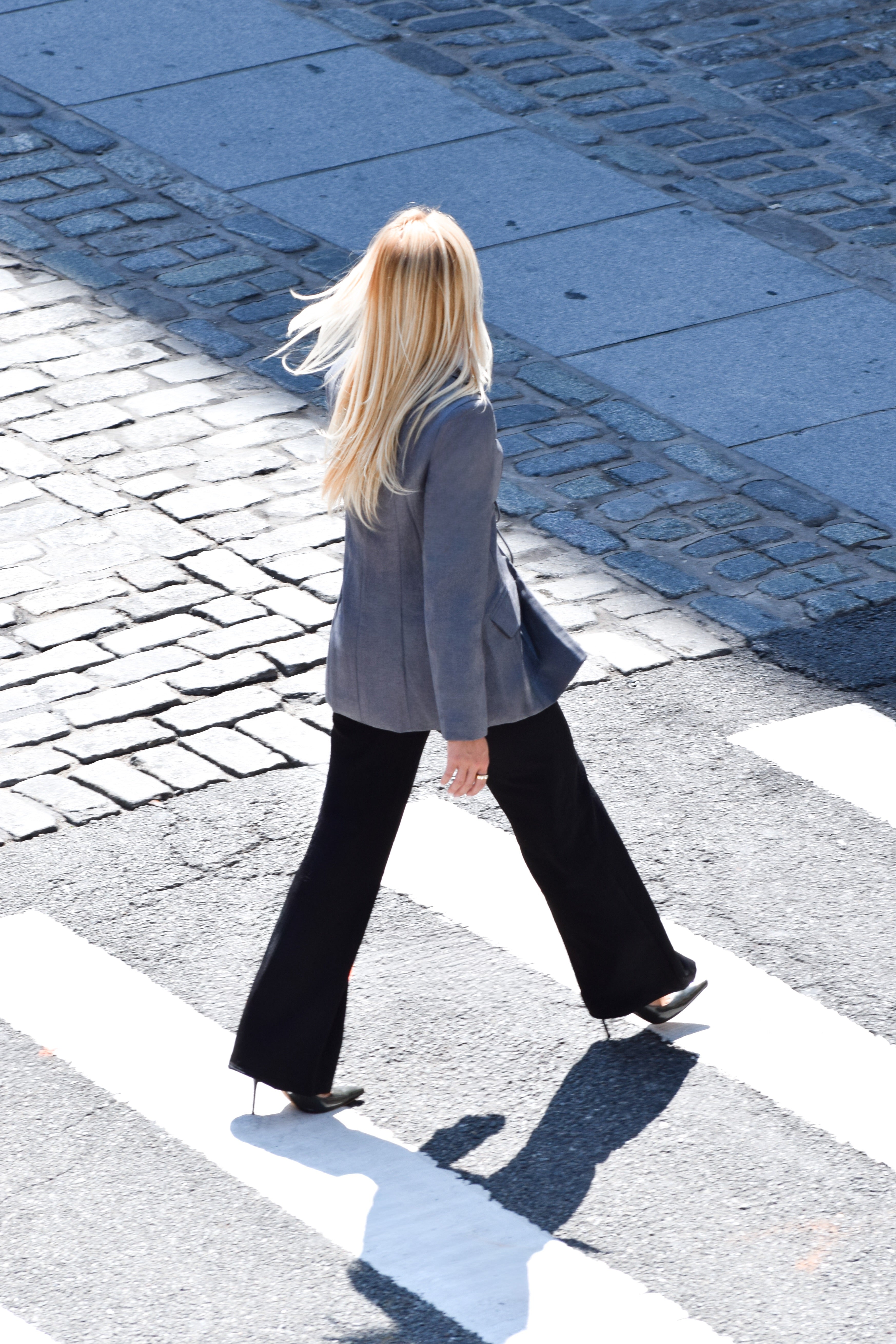 A woman with long blonde hair walks across a zebra crossing from behind, wearing a gray blazer, black flared pants, and black heels on a cobblestone street.