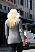 A woman with long blonde hair seen from behind, wearing a structured gray blazer and black pants, walking on a city street with buildings and parked vans in the background.
