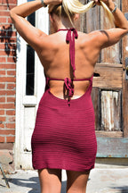 A woman viewed from behind wearing a burgundy knit halter mini dress with cutout back details, standing outdoors in front of a rustic wooden door and brick wall.