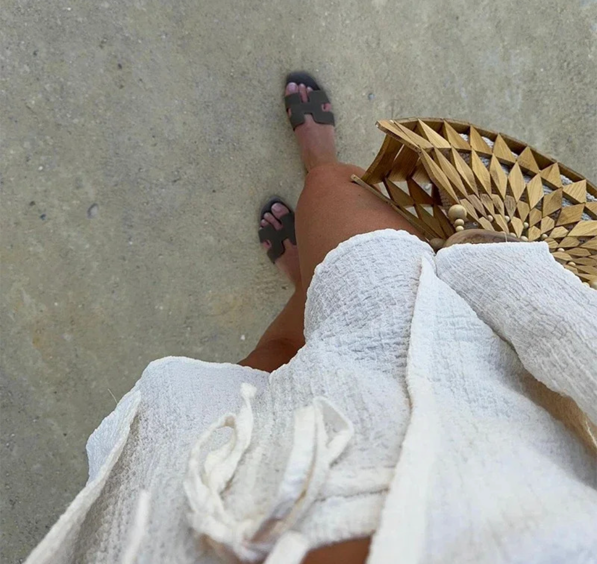 Person wearing a white dress and sandals on a concrete surface with a wicker bag.