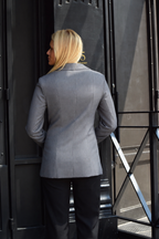 A woman viewed from behind wearing a structured light gray blazer and black trousers, standing in front of a dark doorway.