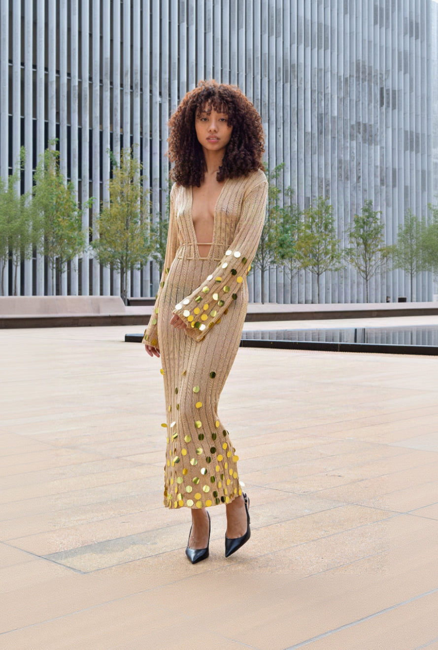 A woman wearing a beige ribbed maxi dress with gold sequin accents, standing outdoors in front of a modern building facade.