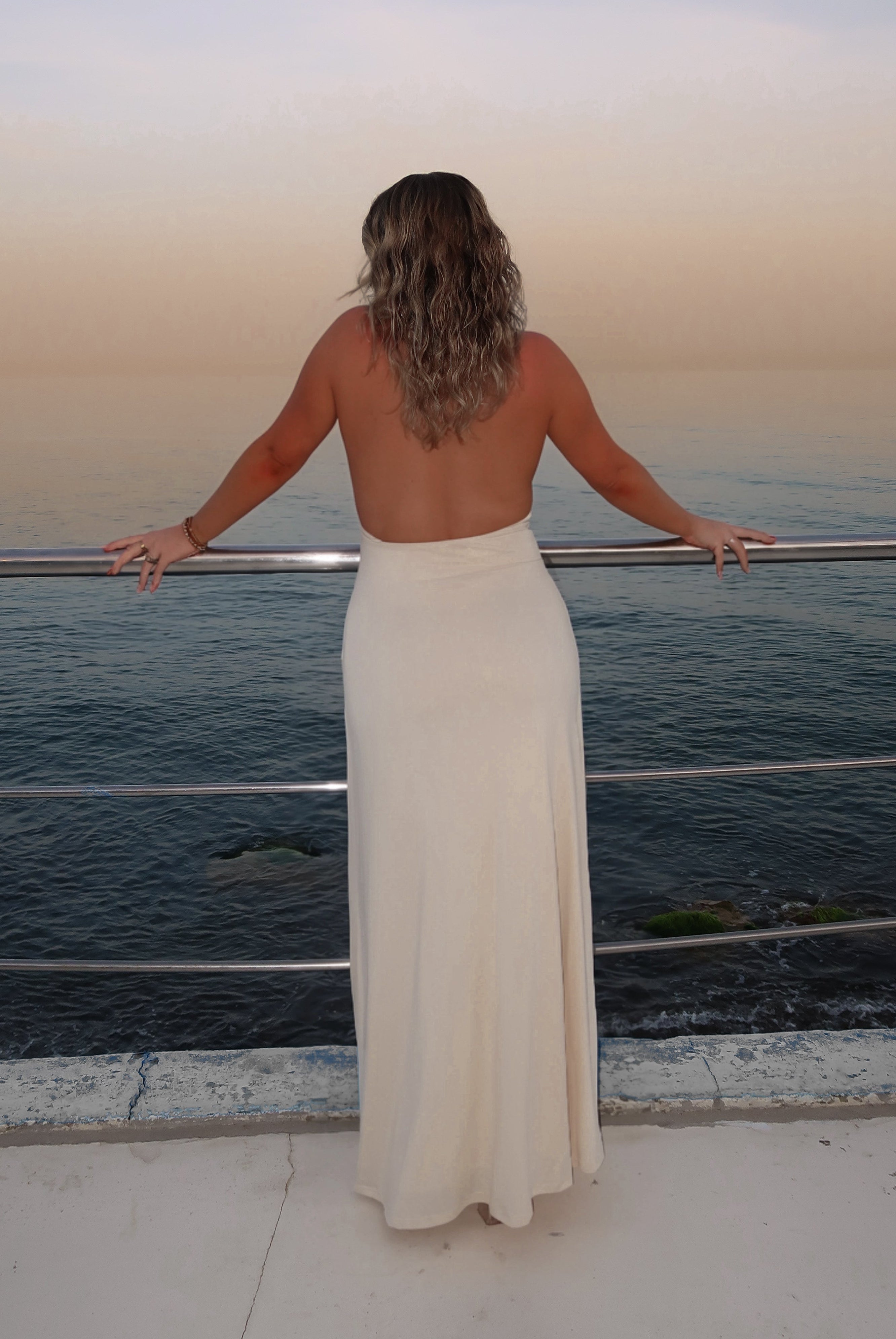 Woman in a white dress standing on a balcony overlooking the ocean at sunset.